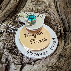 May include: A layered pendant necklace with a silver chain. The pendant features a heart-shaped top piece with a teal gemstone and the date 07-29-21. Below are two circular pieces, one gold and one silver, engraved with "Mayah Flores" and "Forever loved."