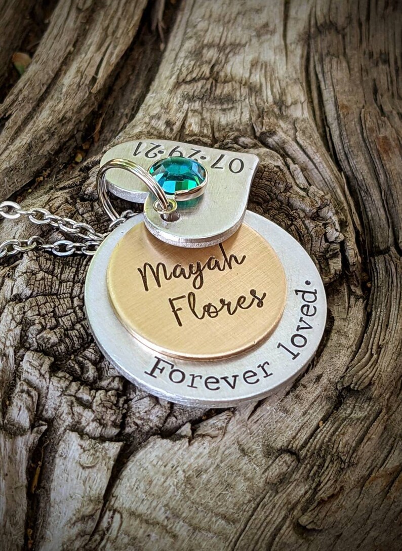 May include: A layered pendant necklace with a silver chain. The pendant features a heart-shaped top piece with a teal gemstone and the date 07-29-21. Below are two circular pieces, one gold and one silver, engraved with "Mayah Flores" and "Forever loved."