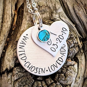 May include: Silver necklace with a round pendant that says "wanted. chosen. loved. added" and a heart pendant that says "03.20.19". The necklace has a teal blue crystal charm.