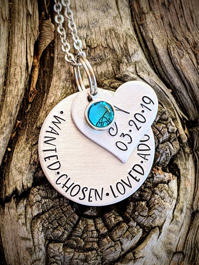 May include: Silver necklace with a round pendant that says "wanted. chosen. loved. added" and a heart pendant that says "03.20.19". The necklace has a teal blue crystal charm.