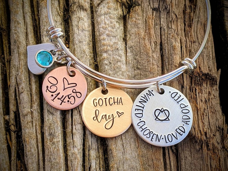 May include: A silver expandable bangle bracelet with three charms. The charms are round and have different engravings. One charm is copper and says "0.3.14.58" with a heart. Another charm is gold and says "GOTCHA day" with a heart. The third charm is silver and says "WANTED. CHOSEN. LOVED. ADOPTED." with a heart and a symbol.