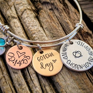 May include: A silver bangle bracelet with three charms. The charms are a copper disc with the date "01.14.58" and a heart, a copper disc with the words "Gotcha Day" and a heart, and a silver disc with the words "Loved. Adopted. Wanted. Chosen." and a heart.