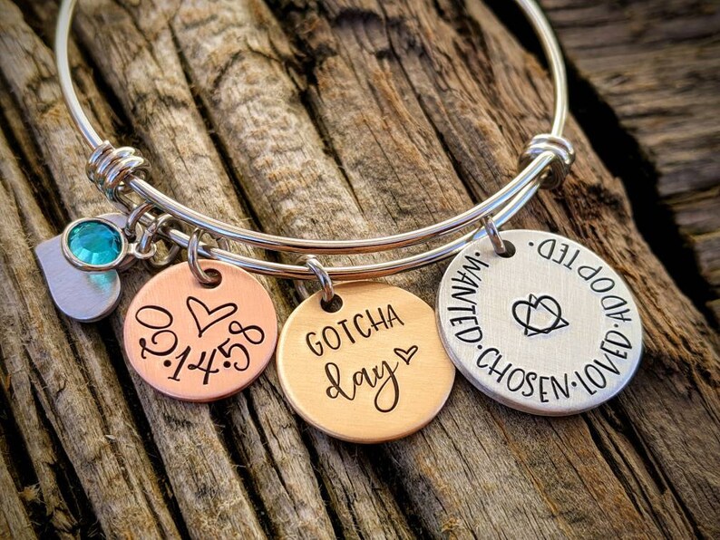 May include: A silver bangle bracelet with three charms. The charms are a copper disc with the date "01.14.58" and a heart, a copper disc with the words "Gotcha Day" and a heart, and a silver disc with the words "Loved. Adopted. Wanted. Chosen." and a heart.