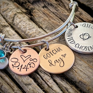 May include: A silver bangle bracelet with three charms. The charms are round and have the words "Gotcha Day", "0.1.14.18", and "Wanted. Chosen. Loved. Adopted" engraved on them. The bracelet also has a small silver heart charm with a turquoise crystal.