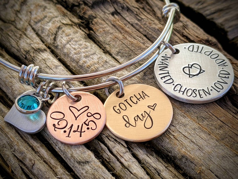 May include: A silver bangle bracelet with three charms. The charms are round and have the words "Gotcha Day", "0.1.14.18", and "Wanted. Chosen. Loved. Adopted" engraved on them. The bracelet also has a small silver heart charm with a turquoise crystal.