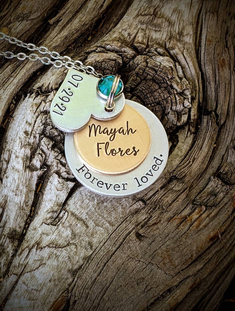 May include: A silver necklace with multiple pendants. The top pendant is heart-shaped with the date "07.29.21" engraved. Below, a round pendant is inscribed with "Mayah Flores" and "Forever loved.". A teal gemstone is set at the top.