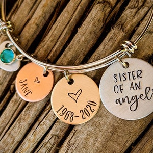 May include: A silver bangle bracelet with charms. The charms include a heart with a green gem, a copper charm with the word "JANE" and a heart, a copper charm with a heart and the dates "1968-2020", and a silver charm with the text "SISTER OF AN angel" and a heart.