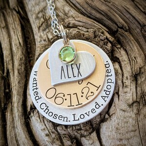 May include: Silver and gold necklace with a heart charm engraved with the name "Alex" and the date "06.11.21". The necklace also has a round charm engraved with the words "Wanted. Chosen. Loved. Adopted."