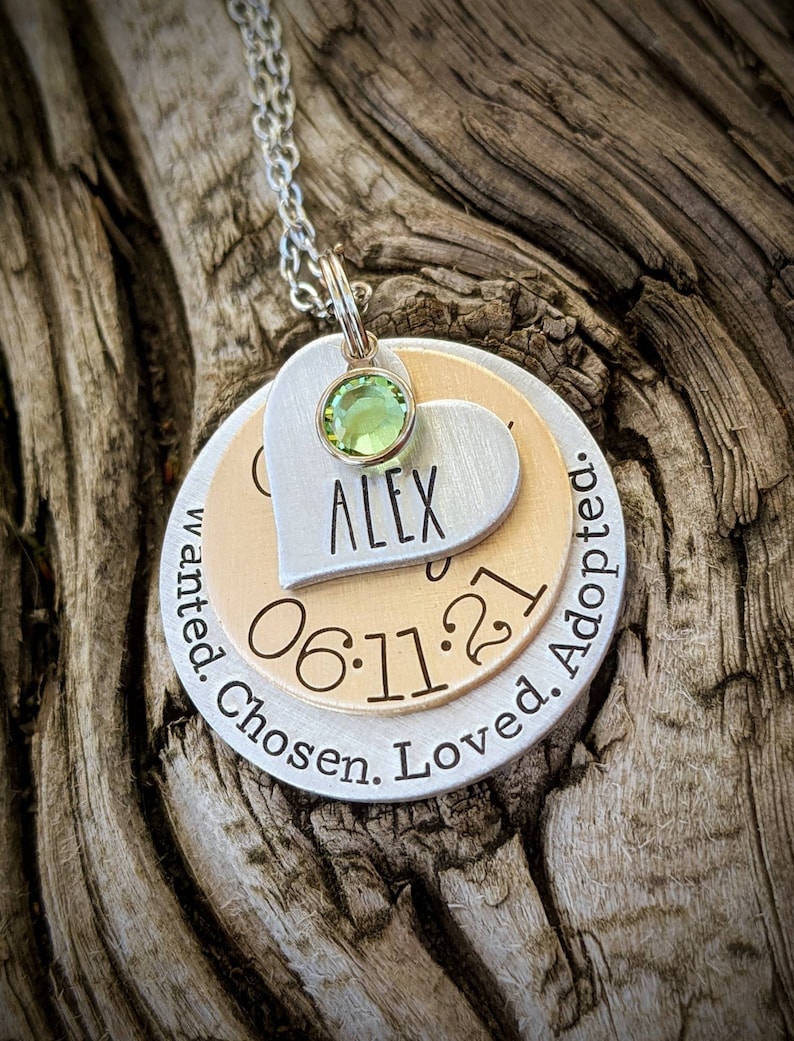 May include: Silver and gold necklace with a heart charm engraved with the name "Alex" and the date "06.11.21". The necklace also has a round charm engraved with the words "Wanted. Chosen. Loved. Adopted."