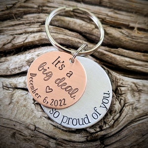 Puede incluir: Un llavero de plata y cobre con el texto "It's a big deal. December 6, 2022" en el lado de cobre y "So proud of you." en el lado de plata.