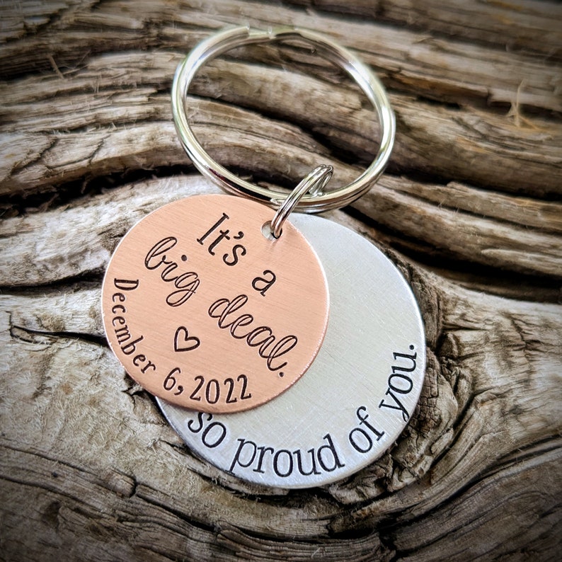 Puede incluir: Un llavero de plata y cobre con el texto "It's a big deal. December 6, 2022" en el lado de cobre y "So proud of you." en el lado de plata.