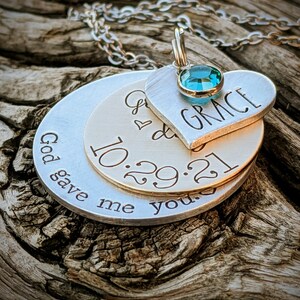May include: Silver necklace with three layered pendants. The top pendant is a heart with the word "GRACE" engraved on it and a turquoise blue gemstone. The middle pendant is a round disc with the date "10.29.21" engraved on it. The bottom pendant is a round disc with the text "God gave me you" engraved on it.