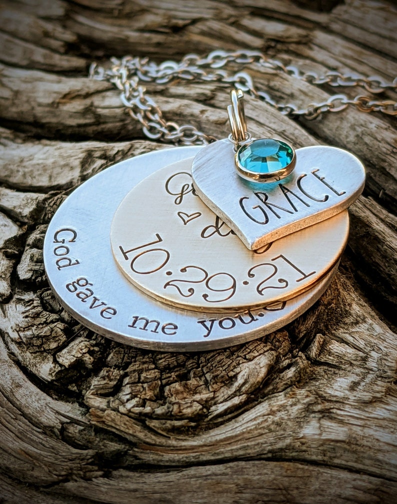 May include: Silver necklace with three layered pendants. The top pendant is a heart with the word "GRACE" engraved on it and a turquoise blue gemstone. The middle pendant is a round disc with the date "10.29.21" engraved on it. The bottom pendant is a round disc with the text "God gave me you" engraved on it.