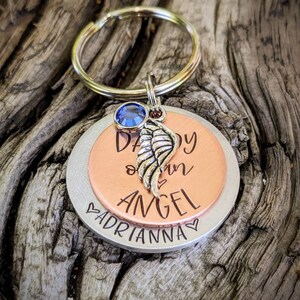 May include: A silver and copper keychain with a blue crystal charm. The keychain has the words "Daddy our Angel" engraved on it, with the name "Adrianna" engraved on the bottom.