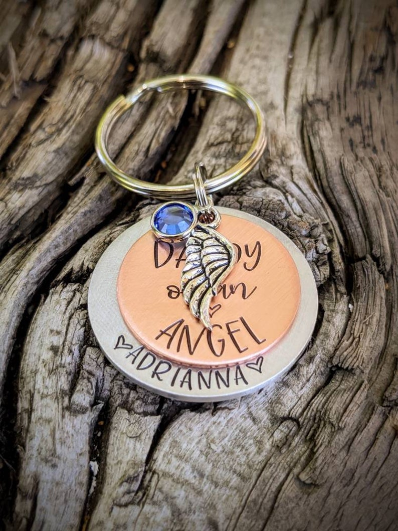 May include: A silver and copper keychain with a blue crystal charm. The keychain has the words "Daddy our Angel" engraved on it, with the name "Adrianna" engraved on the bottom.