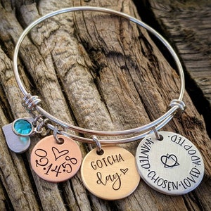 May include: Silver bangle bracelet with three charms. The charms are round and have the following text: "Gotcha Day", "0.1.4.5", and "Chosen. Wanted. Adopted. Loved."