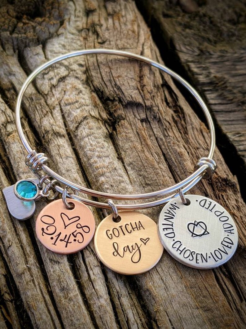 May include: Silver bangle bracelet with three charms. The charms are round and have the following text: "Gotcha Day", "0.1.4.5", and "Chosen. Wanted. Adopted. Loved."