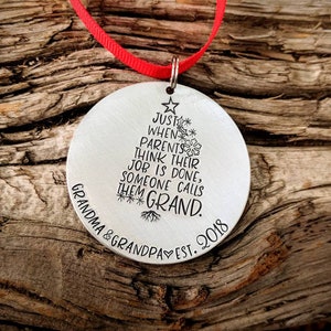 May include: A silver Christmas ornament with a tree design and the text "Just when parents think their job is done, someone calls them Grand. Grandma & Grandpa Est. 2018".