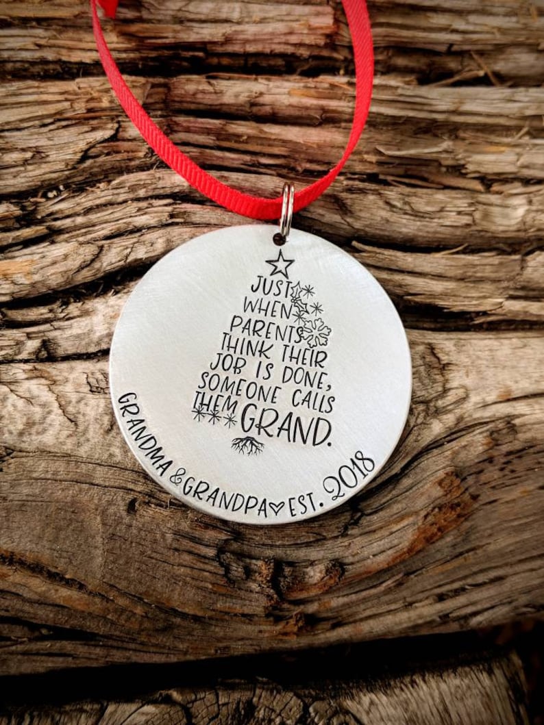 May include: A silver Christmas ornament with a tree design and the text "Just when parents think their job is done, someone calls them Grand. Grandma & Grandpa Est. 2018".