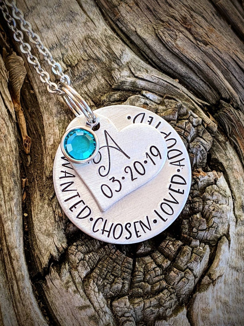 May include: Silver necklace with a round charm that says "Wanted. Chosen. Loved. Addy. 03.2019" and a smaller heart charm with the letter "A" and a turquoise gemstone.