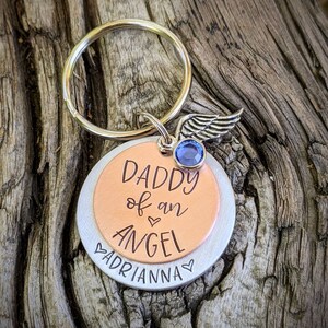 May include: A silver and copper keychain with a blue gem and a wing charm. The keychain has the text "Daddy of an Angel" and "Adrianna" engraved on it.