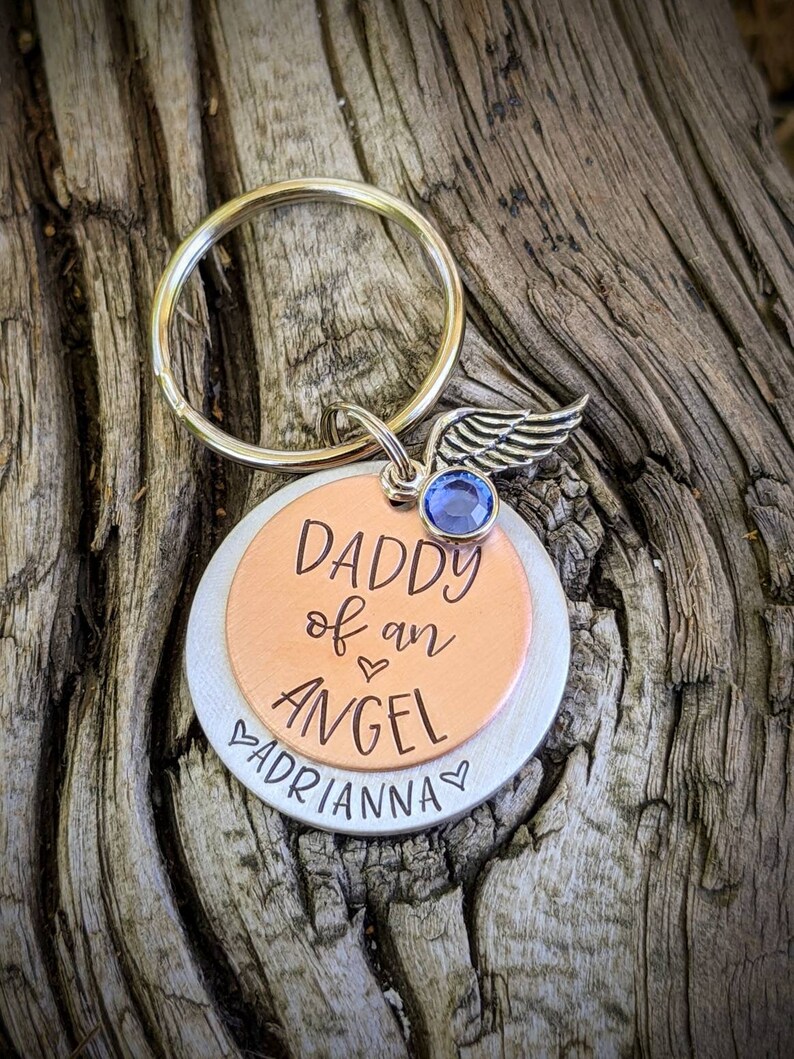 May include: A silver and copper keychain with a blue gem and a wing charm. The keychain has the text "Daddy of an Angel" and "Adrianna" engraved on it.