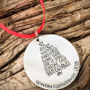 May include: A silver round ornament with a Christmas tree design and the text "Just when parents think their job is done, someone calls them Grand. Grandma & Grandpa est. 2018"