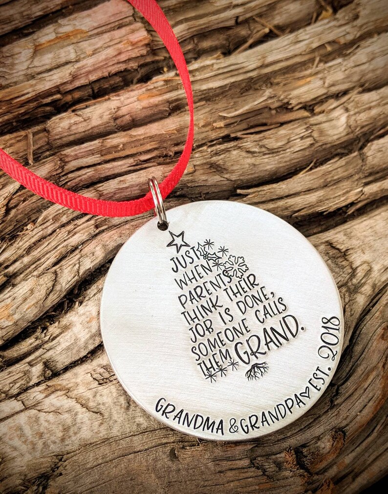 May include: A silver round ornament with a Christmas tree design and the text "Just when parents think their job is done, someone calls them Grand. Grandma & Grandpa est. 2018"