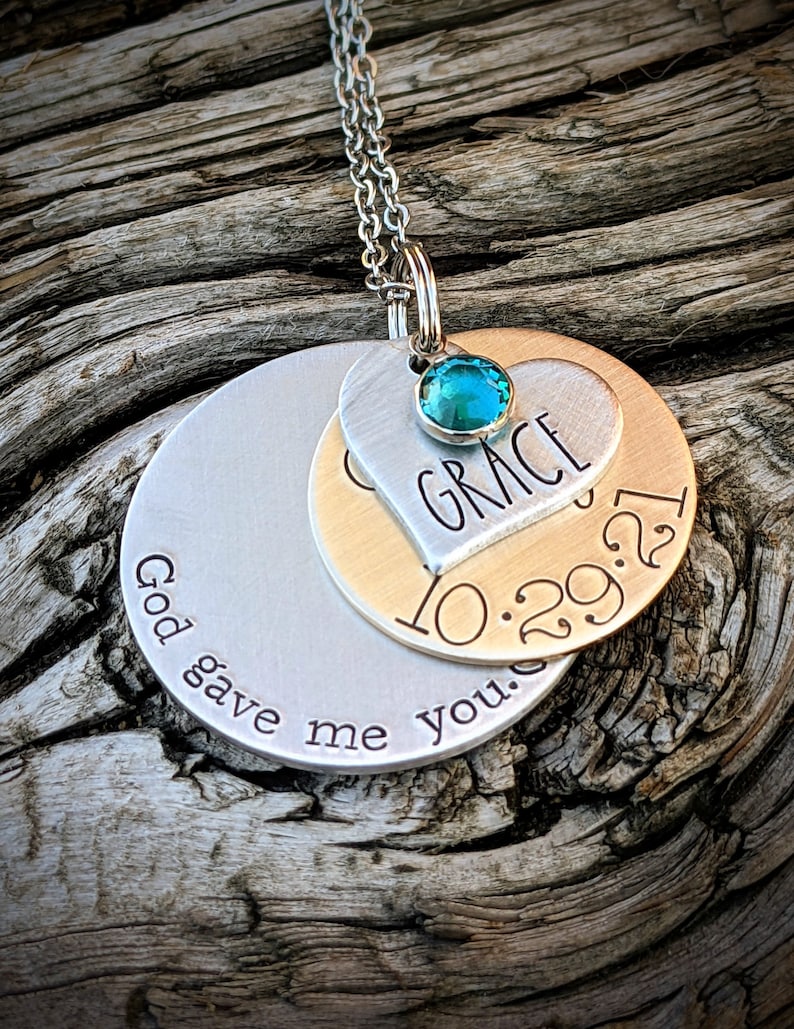 May include: Silver necklace with two round pendants. The top pendant is a heart with the name "GRACE" and the date "10.29.21" engraved on it. The bottom pendant has the text "God gave me you." A turquoise gemstone is set in the heart pendant.