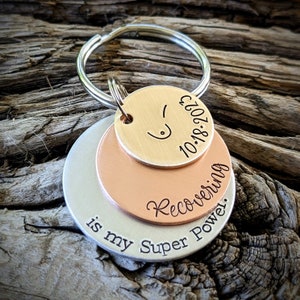 May include: A silver, copper, and gold layered keychain with the text "Recovering is my Super Power." and the date "10.18.2021".