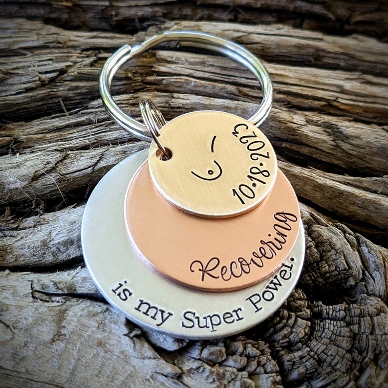 May include: A silver, copper, and gold layered keychain with the text "Recovering is my Super Power." and the date "10.18.2021".