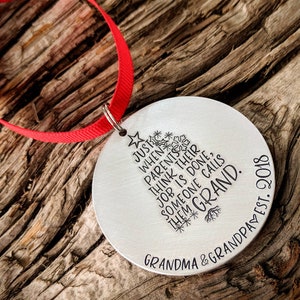 May include: A silver round Christmas ornament with a red ribbon. The ornament has a stylized tree design with the text "JUST WHEN PARENTS THINK THEIR JOB IS DONE, SOMEONE CALLS THEM GRAND." and "GRANDMA & GRANDPA EST. 2018".