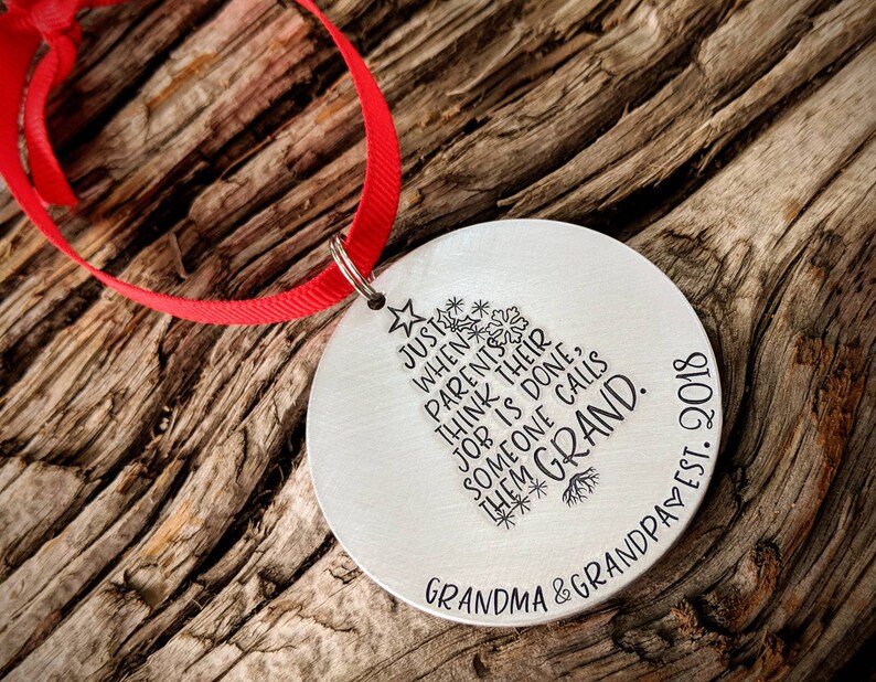 May include: A silver round Christmas ornament with a red ribbon. The ornament has a stylized tree design with the text "JUST WHEN PARENTS THINK THEIR JOB IS DONE, SOMEONE CALLS THEM GRAND." and "GRANDMA & GRANDPA EST. 2018".