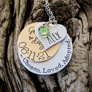May include: Silver and gold necklace with a heart charm engraved with the name "Alex" and the date "06.11.21". The necklace also has a round charm with the text "Chosen. Loved. Adopted."