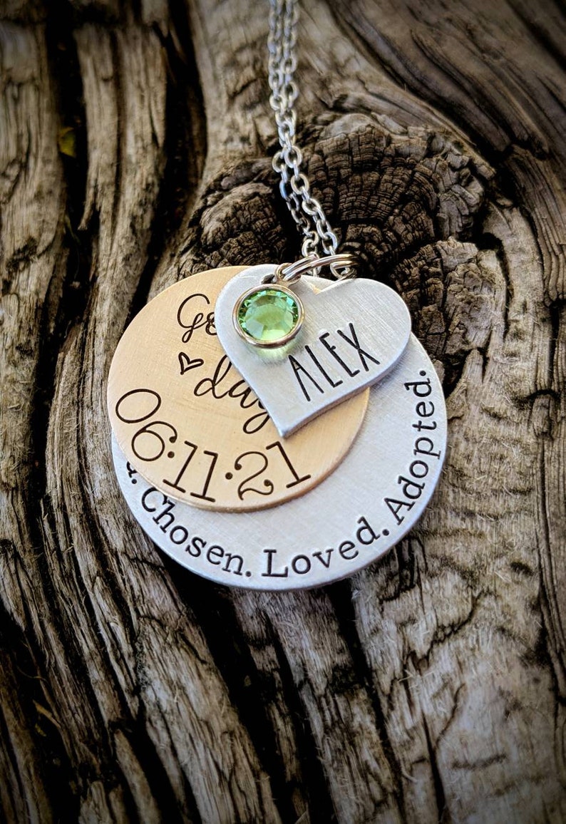 May include: Silver and gold necklace with a heart charm engraved with the name "Alex" and the date "06.11.21". The necklace also has a round charm with the text "Chosen. Loved. Adopted."