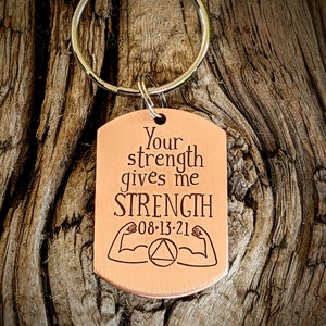 May include: A copper-colored dog tag keychain with the words "Your strength gives me STRENGTH" engraved in black. Below the text are two flexing arm illustrations and the date "08-13-21". The keychain has a silver ring.