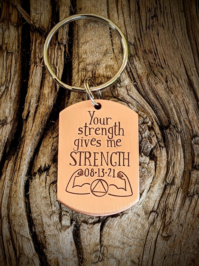 May include: A copper-colored dog tag keychain with the words "Your strength gives me STRENGTH" engraved in black. Below the text are two flexing arm illustrations and the date "08-13-21". The keychain has a silver ring.