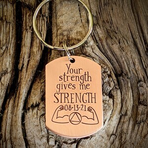 May include: A copper-colored dog tag keychain with the text "Your strength gives me STRENGTH" and the date "08-13-21". The tag also features illustrations of muscular arms and a triangle symbol. The keychain ring is silver.