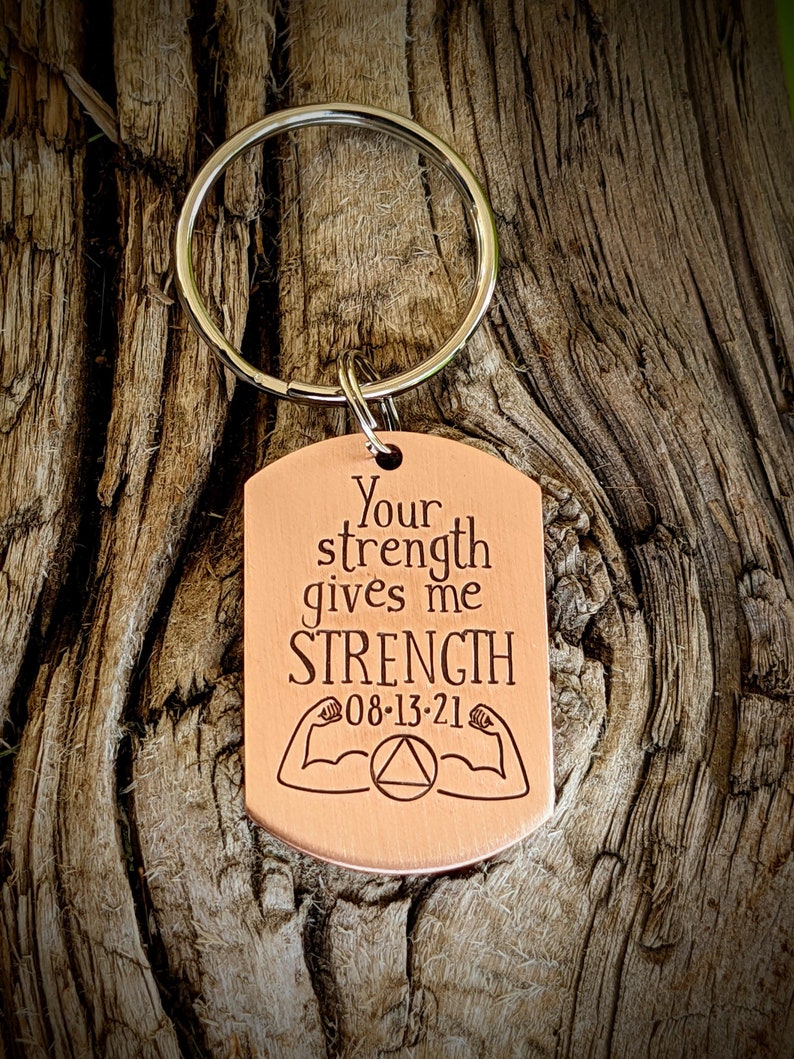 May include: A copper-colored dog tag keychain with the text "Your strength gives me STRENGTH" and the date "08-13-21". The tag also features illustrations of muscular arms and a triangle symbol. The keychain ring is silver.