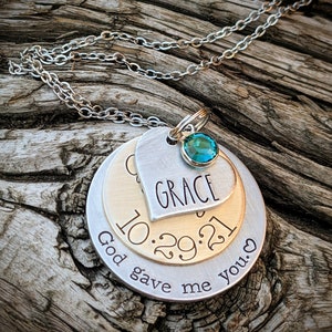 May include: A silver chain necklace with a layered pendant. The pendant features a heart with the word "GRACE" inscribed, a round disc with the date "10.29.21", and a larger round disc with the text "God gave me you." A teal blue gemstone is attached to the heart pendant.