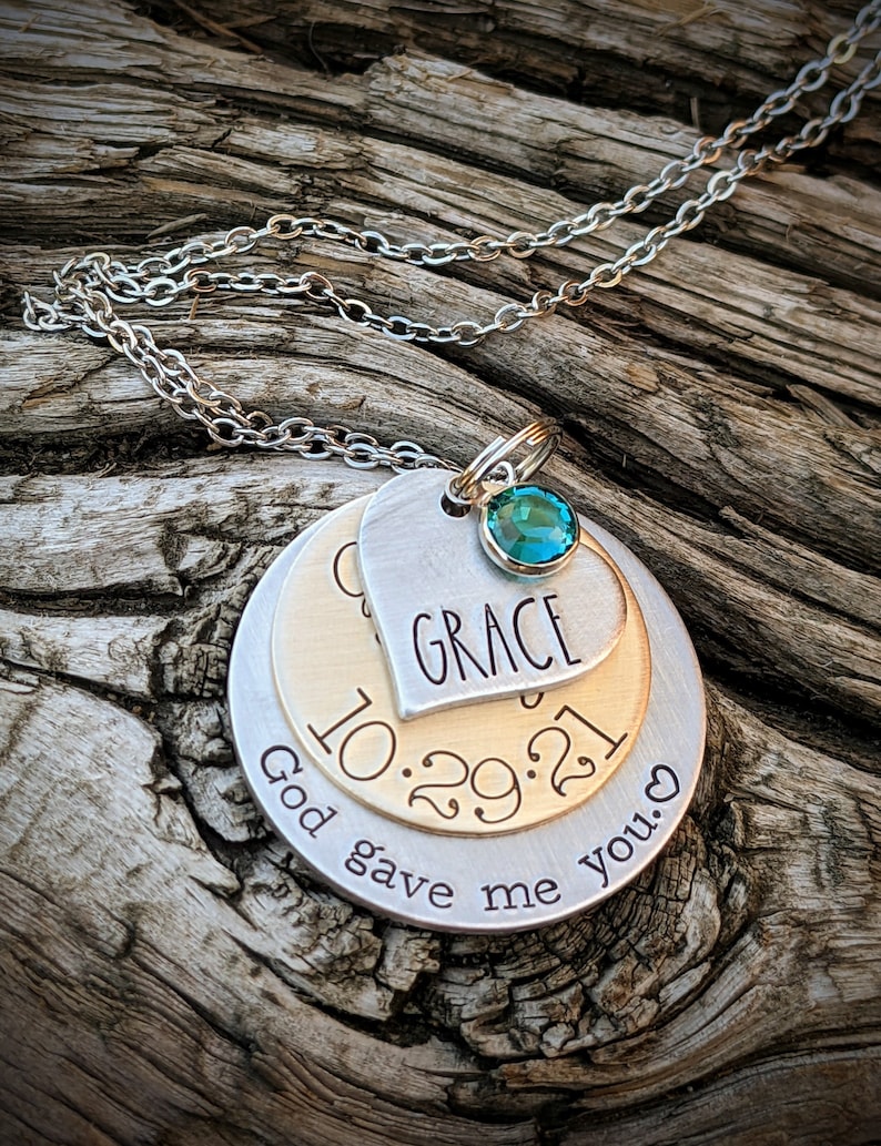 May include: A silver chain necklace with a layered pendant. The pendant features a heart with the word "GRACE" inscribed, a round disc with the date "10.29.21", and a larger round disc with the text "God gave me you." A teal blue gemstone is attached to the heart pendant.