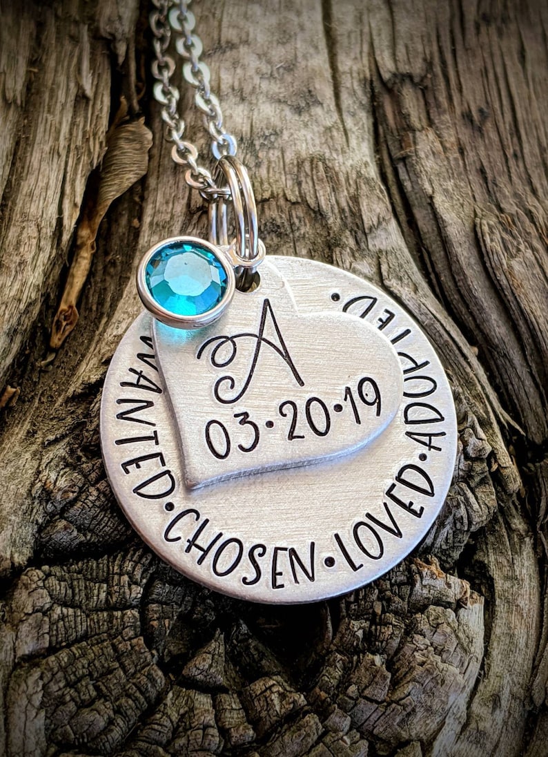 May include: Silver necklace with a heart charm engraved with the letter "A", the date "03.20.19", and the words "wanted, chosen, loved, adopted". A small turquoise gemstone dangles from the chain.