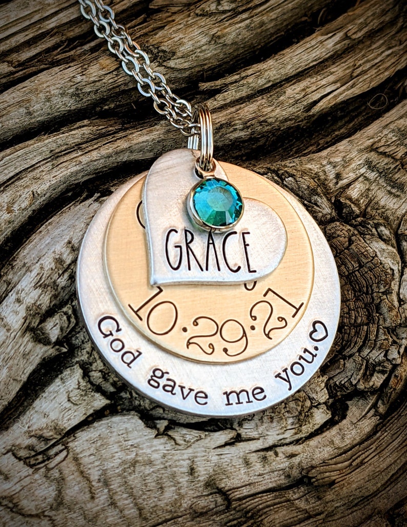 May include: A silver necklace with a layered pendant featuring a heart with the word "GRACE" and the date "10.29.21" on a gold disc. The pendant also includes a teal gemstone and the text "God gave me you."