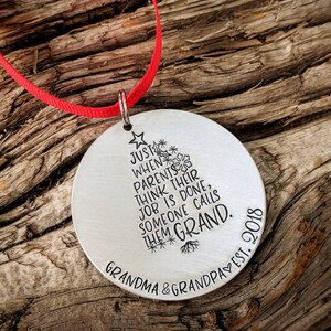 May include: Silver Christmas ornament with a stylized tree design and the text "JUST WHEN PARENTS THINK THEIR JOB IS DONE, SOMEONE CALLS THEM GRAND. GRANDMA & GRANDPA EST. 2018"