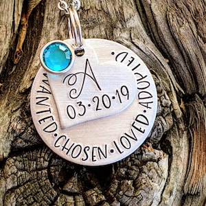 May include: Silver necklace with a round pendant that says "Wanted. Chosen. Loved. Adopted." and a heart charm with the letter "A" and the date "03.20.19". A turquoise gemstone is attached to the necklace.
