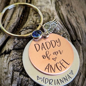 May include: A silver and copper keychain with a blue crystal charm and a silver angel wing charm. The keychain has the text "Daddy of an Angel" on the copper side and "Adrianna" on the silver side.