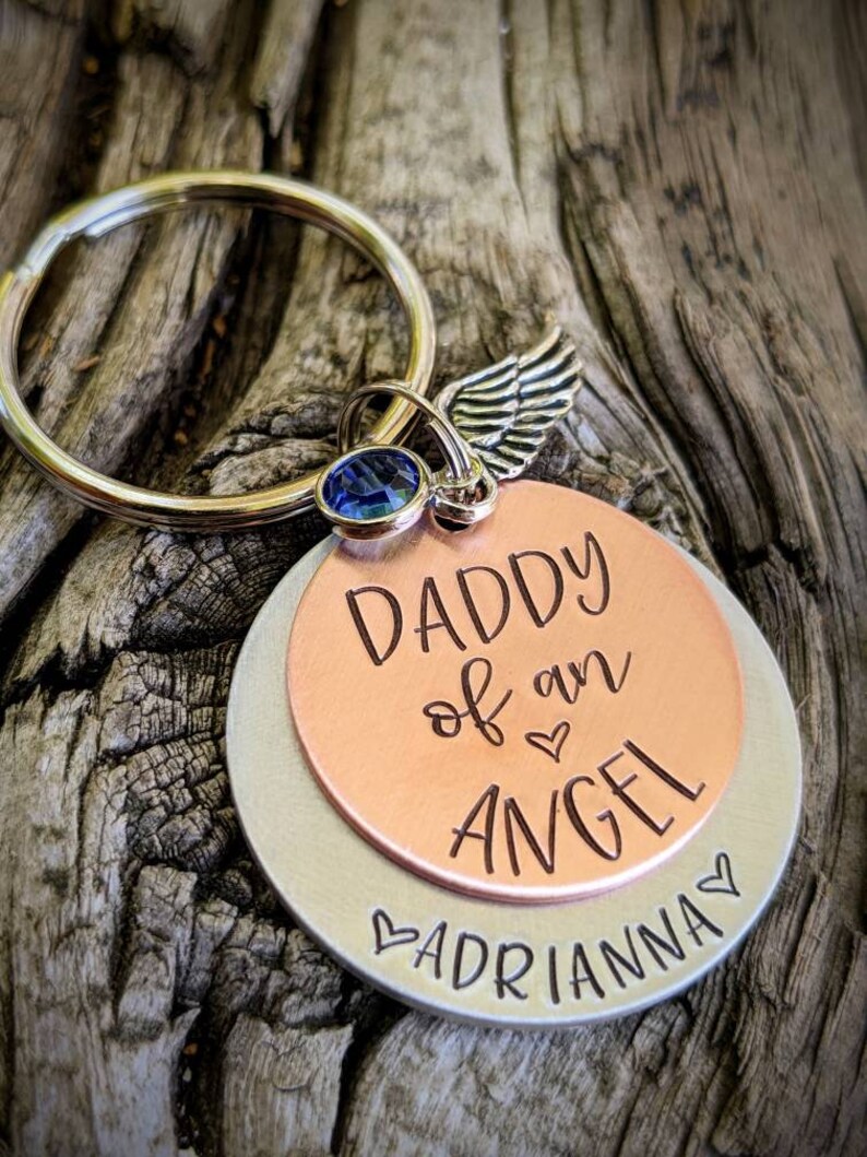 May include: A silver and copper keychain with a blue crystal charm and a silver angel wing charm. The keychain has the text "Daddy of an Angel" on the copper side and "Adrianna" on the silver side.
