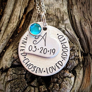 May include: Silver necklace with a heart charm that says "Chosen. Loved. Adopted." and the date "03.20.19". The heart charm has a small turquoise gemstone attached to it.