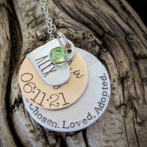May include: Silver necklace with three layered discs. The top disc is a heart with the name "Alex" engraved on it and a green gemstone. The middle disc is gold and has the date "06.11.21" engraved on it. The bottom disc is silver and has the text "Chosen. Loved. Adopted." engraved on it.