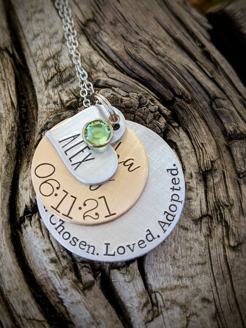 May include: Silver necklace with three layered discs. The top disc is a heart with the name "Alex" engraved on it and a green gemstone. The middle disc is gold and has the date "06.11.21" engraved on it. The bottom disc is silver and has the text "Chosen. Loved. Adopted." engraved on it.